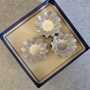 May include: Three silver metal baking molds in the shape of a flower with fluted edges. The molds are in a blue box with a white interior.