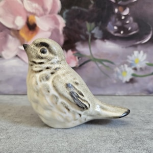May include: A small ceramic bird figurine featuring a light beige and cream base color with dark brown and gray speckled markings on the head, wings, and chest. The bird is depicted in a perched position with a pointed beak and a smooth finish.