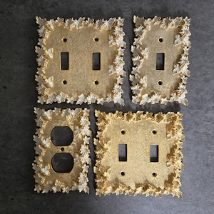 May include: Four decorative light switch covers with a gold finish and leaf-like trim. The covers include single and double switch configurations, as well as an outlet cover. The leaf trim is a lighter gold color.