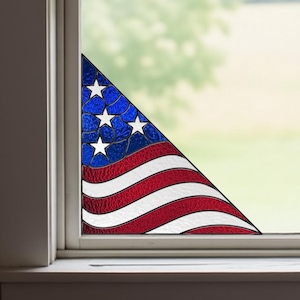 Pode incluir: Uma moldura de janela branca com um canto de um desenho da bandeira americana. A bandeira apresenta um campo azul com estrelas brancas e listras vermelhas e brancas. A janela tem vista para um campo verde e árvores.