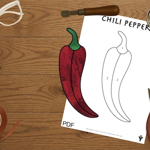 May include: A sheet with a chili pepper design, labeled with the text "CHILI PEPPER" and numbered sections. Tools like pliers, a tape measure, and a glass cutter are arranged around the sheet on a wooden surface.