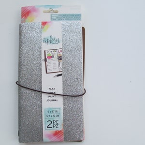 May include: A silver glitter-covered journal with a dark brown elastic band. The paper belly band features the text The Explorer Journal, Plan Draw Paint Journal, and 5 x 8.7 in. The packaging includes a header card with watercolor accents.