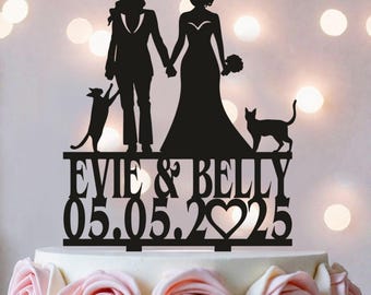 May include: A black cake topper featuring silhouettes of two people holding hands, accompanied by two cats. The text reads EVIE & BELLY above the date 05.05.2025, with a heart shape replacing the zero in the year, all standing on a cake base.