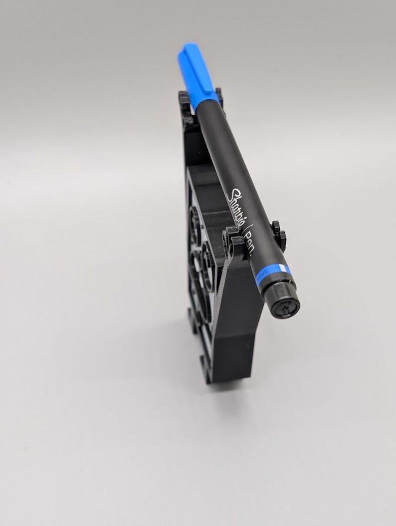 Puede incluir: Un portal&aacute;pices negro con un bol&iacute;grafo Sharpie azul. El bol&iacute;grafo es negro con detalles azules y el texto "Sharpie Pen" impreso en el lateral. El portal&aacute;pices es negro y tiene un dise&ntilde;o geom&eacute;trico.