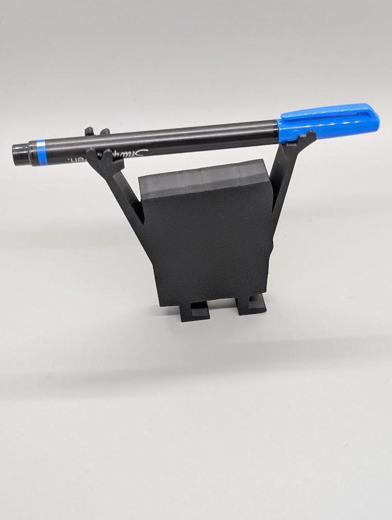 Puede incluir: Un portal&aacute;pices negro con un bol&iacute;grafo negro y una tapa azul. El bol&iacute;grafo descansa sobre un soporte negro de forma geom&eacute;trica. El bol&iacute;grafo tiene la palabra "Sharpie" impresa.