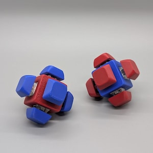 Puede incluir: Dos rompecabezas geométricos, cada uno con un cubo central rojo y piezas de esquina azules. Los rompecabezas tienen un diseño simétrico con colores rojo y azul. El fondo es gris liso.