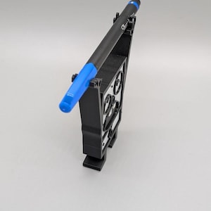 Puede incluir: Un bol&iacute;grafo negro con una tapa azul descansa en un soporte negro para bol&iacute;grafos. El soporte para bol&iacute;grafos tiene un dise&ntilde;o geom&eacute;trico con formas recortadas. El bol&iacute;grafo est&aacute; en &aacute;ngulo, mostrando su punta azul y la marca Sharpie.