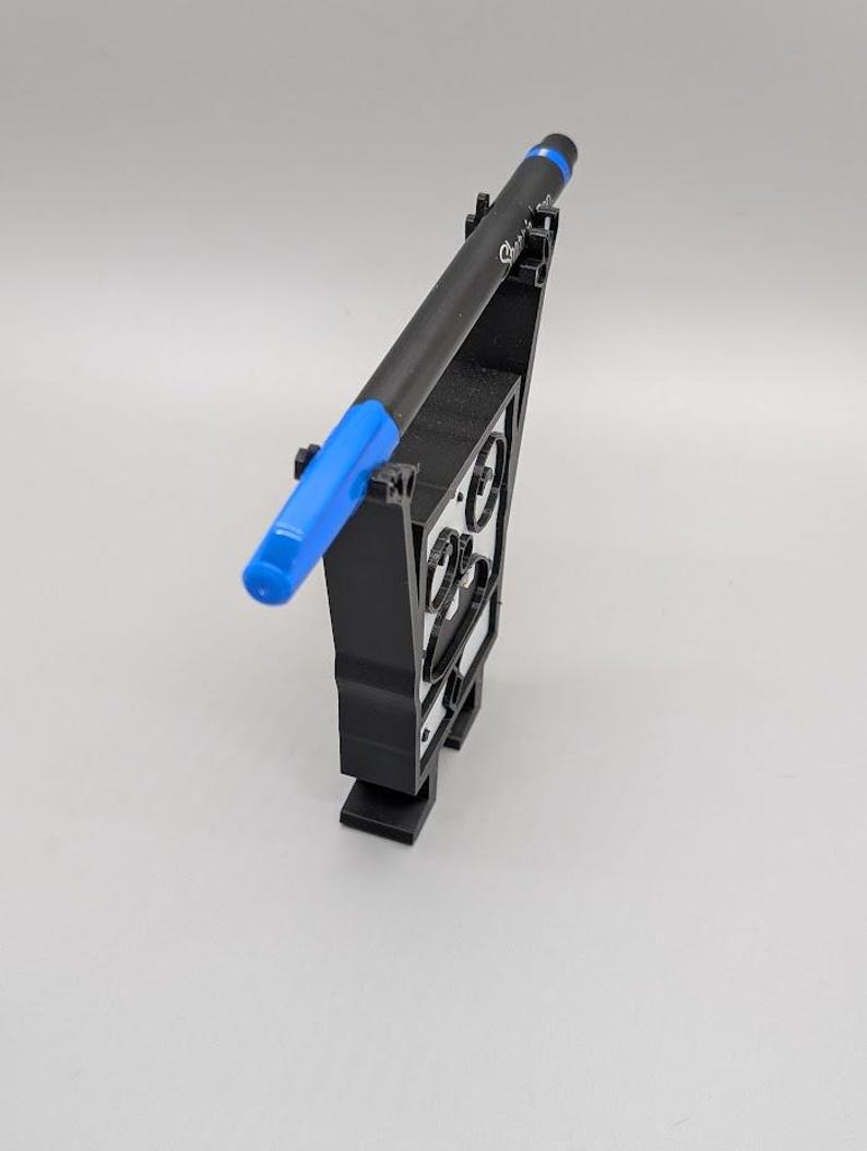 Puede incluir: Un bol&iacute;grafo negro con una tapa azul descansa en un soporte negro para bol&iacute;grafos. El soporte para bol&iacute;grafos tiene un dise&ntilde;o geom&eacute;trico con formas recortadas. El bol&iacute;grafo est&aacute; en &aacute;ngulo, mostrando su punta azul y la marca Sharpie.