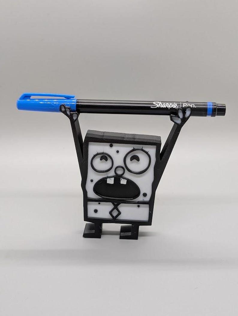 Puede incluir: Una figura de Bob Esponja en blanco y negro impresa en 3D sosteniendo un bol&iacute;grafo Sharpie negro con una tapa azul. La figura tiene una expresi&oacute;n de sorpresa. El bol&iacute;grafo mide aproximadamente 14 cm de largo. La figura est&aacute; sobre una superficie plana.