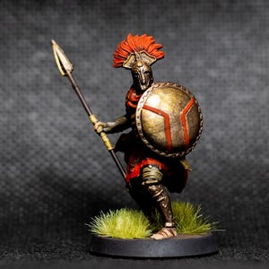 May include: Miniature Spartan warrior figurine, holding a spear and shield. The warrior wears a red tunic, bronze armor, and a crested helmet. The shield is gold with a red design. The figurine stands on a small, round base with artificial grass.