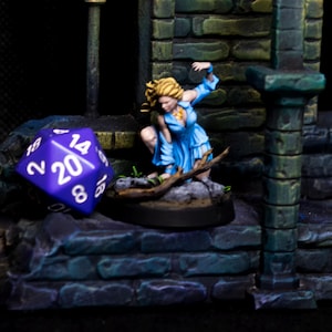 May include: A miniature fantasy figure of a woman in a blue dress, posed in a dungeon setting. A large purple twenty-sided die rests nearby. The scene is detailed with stone textures and a dark, dramatic atmosphere.