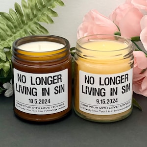 Könnte beinhalten: Zwei Glas-Kerzengläser mit weißen Etiketten mit der Aufschrift „NO LONGER LIVING IN SIN“ und Datumsangaben. Eine Kerze befindet sich in einem braunen Glas mit weißem Wachs, die andere in einem klaren Glas mit gelbem Wachs. Der Hintergrund umfasst Grün und rosa Blumen.