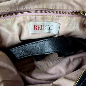 May include: Inside view of a black handbag with a black leather strap. The lining is a light pink fabric. A white tag with the text "RED V" in red is visible. The zipper is gold-coloured.