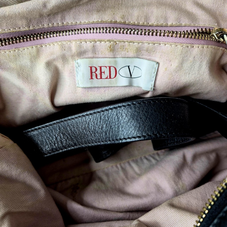 May include: Inside view of a black handbag with a black leather strap. The lining is a light pink fabric. A white tag with the text "RED V" in red is visible. The zipper is gold-coloured.