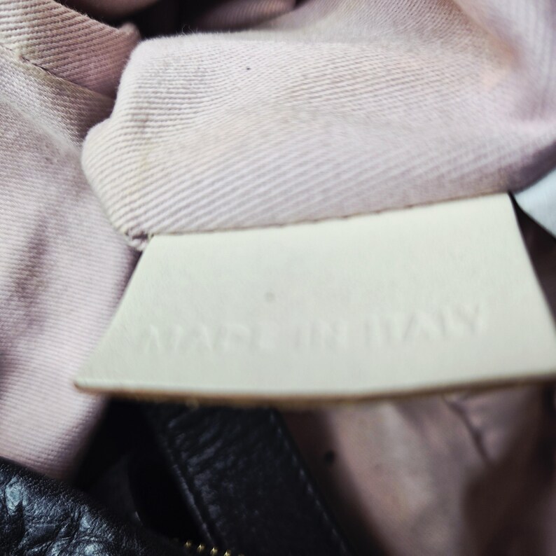 May include: Close-up of a white leather tag with the embossed text "MADE IN ITALY". The tag is sewn into a light pink fabric lining, with a black leather material visible at the bottom of the image.