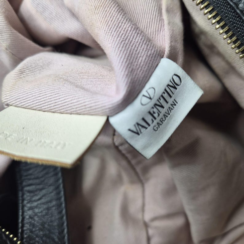May include: Close-up of a black leather bag interior with a light pink fabric lining. A white tag with the "VALENTINO GARAVANI" logo is visible, along with a smaller tag that reads "MADE IN ITALY".