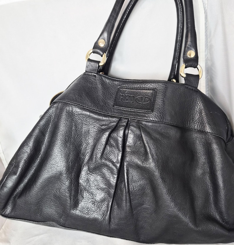 May include: Black leather handbag with gold-coloured hardware. The bag has a pleated design on the front and a top zip closure. The brand name "RED VALENTINO" is embossed on a leather patch.