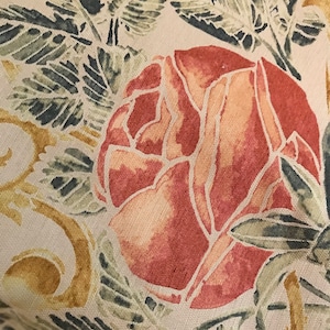 May include: Close-up of a floral fabric featuring a large, stylized rose in shades of peach and red. The rose is surrounded by green and yellow leaves and stems on a cream-colored background. The design has a watercolor-like quality.