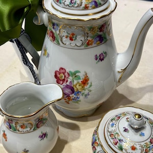 May include: A white porcelain tea set with floral designs and gold trim. The set includes a teapot, creamer, and sugar bowl. A green ribbon is tied to the handle of the teapot. The floral pattern features red, purple, yellow, and blue flowers.