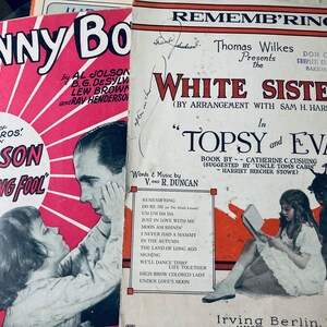 Könnte beinhalten: Zwei Vintage-Notenblätter. Das linke Cover ist rosa mit einer Schwarzweißillustration von Al Jolson im Musical "The Singing Fool". Das rechte Cover ist rot mit einer Schwarzweißillustration von zwei Kindern aus dem Musical "Topsy and Eva".