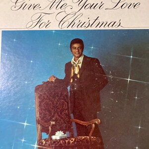 May include: A vintage vinyl record album cover featuring a man in a suit standing next to a chair. The album cover is blue with white stars and the text "Johnny Mathis Give Me Your Love For Christmas".