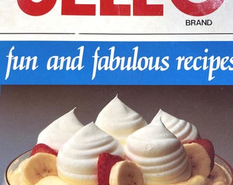 Op de afbeelding: Een vintage kookboek van het merk Jell-O met de tekst "fun and fabulous recipes" op de voorkant. De voorkant toont een foto van een dessert met slagroom, bananen en aardbeien in een glazen kom.