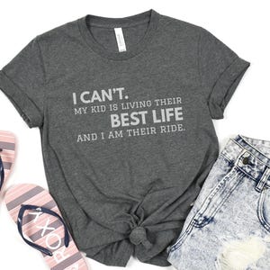 May include: Dark gray t-shirt with the text "I CAN'T. MY KID IS LIVING THEIR BEST LIFE AND I AM THEIR RIDE." The shirt is paired with denim shorts and pink and white striped flip-flops.