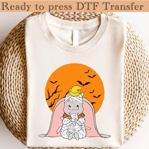 May include: Cream-colored t-shirt with a Halloween-themed graphic. The graphic features a cartoon elephant wrapped in bandages, wearing a yellow hat, set against an orange sun with bats and branches. The text "Ready to press DTF Transfer" is at the top.