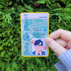 Puede incluir: Una tarjeta de identificación de empleado del Daily Planet con el texto "Daily Planet" y "Clark Kent, periodista". La tarjeta tiene un borde amarillo e incluye un retrato de Clark Kent con gafas. También hay corazones y estrellas decorativas.