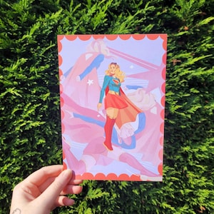 May include: A rectangular art print featuring a central illustration of a blonde superheroine wearing a blue long-sleeved top with a red and yellow emblem, a red skirt, and red boots, surrounded by translucent, layered silhouettes of the same figure.
