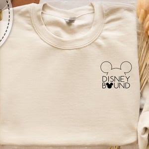 May include: Cream-colored sweatshirt with a crew neck. The design features a black outline of Mickey Mouse ears above the words "DISNEY BOUND" in black. The sweatshirt is a neutral color.