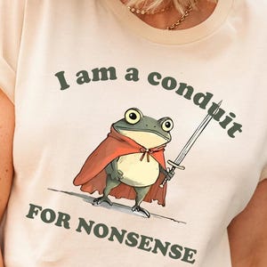 May include: Beige t-shirt with the text "I am a conduit for nonsense." The graphic features a cartoon frog wearing a red cape and holding a sword. The frog has large green eyes and a white belly.
