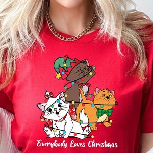 May include: Red t-shirt with a Christmas tree design featuring three cartoon cats. The cats are decorated with Christmas lights and the text "Everybody Loves Christmas" is printed below the design.