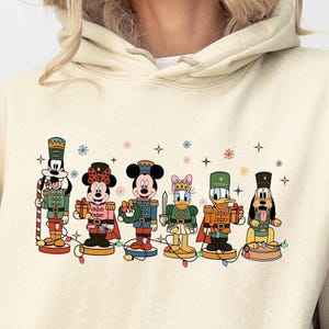 May include: Cream-colored hoodie featuring a festive graphic of Disney characters as nutcrackers. The characters include Goofy, Minnie Mouse, Mickey Mouse, Daisy Duck, Donald Duck, and Pluto, all in holiday attire. Colorful snowflakes and stars surround the characters.