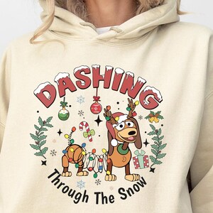 K&ouml;nnte beinhalten: Beige Kapuzenpullover mit einem weihnachtlichen Motiv. Das Design zeigt einen Cartoon-Hund mit Geweih und Lichterketten, mit dem Text "Dashing Through The Snow". Festliche Ornamente und eine Geschenkbox sind ebenfalls vorhanden.