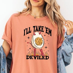 以下が含まれることがあります： コーラル色のTシャツに、角と尻尾のある悪魔の卵のグラフィックがプリントされています。Tシャツには「I'll Take 'Em Deviled」という文字がプリントされています。