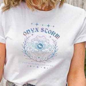 K&ouml;nnte beinhalten: Wei&szlig;es T-Shirt mit einem grafischen Design, das ein Blumenmuster und den Text "ONYX STORM" zeigt. Das Design ist in Blaut&ouml;nen, Rosa und Lila gehalten.