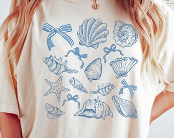 May include: Cream-colored t-shirt featuring a collection of blue seashell illustrations and bows. The design includes various shell shapes and starfish. A casual, beach-themed top.
