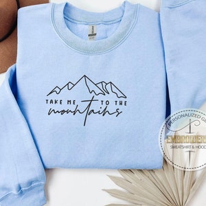 K&ouml;nnte beinhalten: Hellblaues Sweatshirt mit dem schwarzen Text "TAKE ME TO THE mountains". Das Design zeigt eine Bergkette. Im Hintergrund ist ein brauner Hut zu sehen.