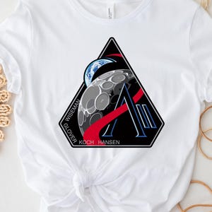 May include: White t-shirt featuring a black, red, and blue graphic design. The design includes a stylized letter "A" with a moon and Earth illustration, and the names "WISEMAN GLOVER KOCH HANSEN" around the edges. A casual, crew neck tee.