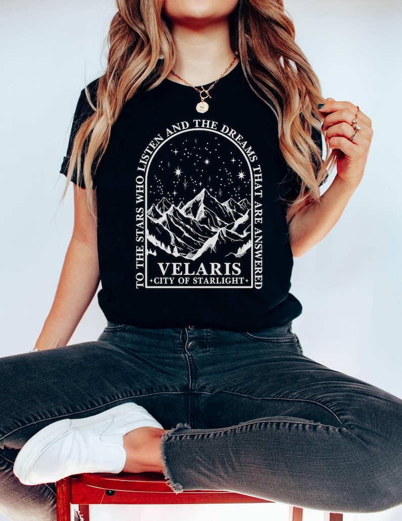 May include: Black t-shirt with a white graphic featuring a mountain range under a starry sky, framed by an arch. Text reads "TO THE STARS WHO LISTEN AND THE DREAMS THAT ARE ANSWERED" and "VELARIS CITY OF STARLIGHT."