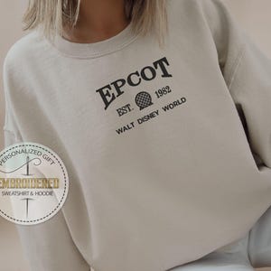 May include: A beige sweatshirt featuring the word "EPCOT" in black, with "EST. 1982" and a sphere graphic. The text "WALT DISNEY WORLD" is below. A circular logo with text "PERSONALIZED GIFT EMBROIDERED SWEATSHIRT & HOODIE" is on the left.