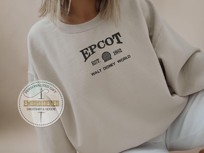 May include: A beige sweatshirt featuring the word "EPCOT" in black, with "EST. 1982" and a sphere graphic. The text "WALT DISNEY WORLD" is below. A circular logo with text "PERSONALIZED GIFT EMBROIDERED SWEATSHIRT & HOODIE" is on the left.