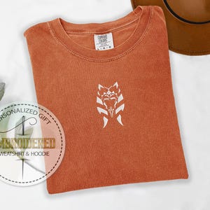 May include: A burnt orange t-shirt with a white embroidered design of a Star Wars character's helmet. The text "Personalized Gift" and "Embroidered Sweatshirt & Hoodie" is on a white circle.
