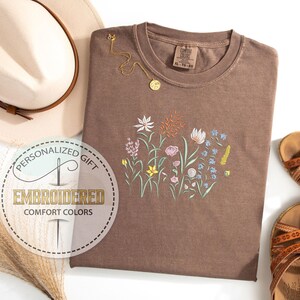 May include: A brown t-shirt with an embroidered floral design. The design features a variety of flowers in shades of pink, blue, yellow, and green. The t-shirt is a comfortable, casual style.