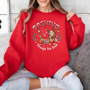 K&ouml;nnte beinhalten: Ein rotes Sweatshirt mit dem Text "Dashing Through The Snow" in einem kreisf&ouml;rmigen Design. Das Design zeigt einen Cartoon-Hund mit Rentiergeweih und einen Dackel, der mit Lichterketten geschm&uuml;ckt ist. Das Sweatshirt ist ein festlicher Weihnachtsartikel.