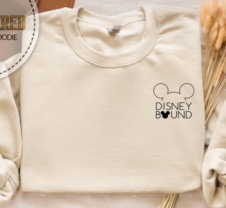 May include: Cream-colored sweatshirt with a crew neck. The design features a black outline of Mickey Mouse ears above the words "DISNEY BOUND" in black. The sweatshirt is a neutral color.