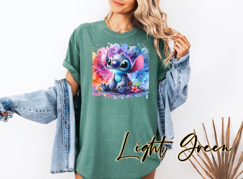 May include: Light green t-shirt with a watercolour illustration of Stitch from Lilo & Stitch. The artwork shows Stitch with a colourful background. The words "Light Green" are written in cursive on the shirt.