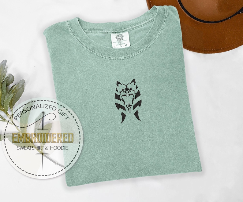 May include: A light green t-shirt with a black embroidered design of a Star Wars character's helmet. The text "Personalized Gift" and "Embroidered Sweatshirt & Hoodie" is printed on a white circle.