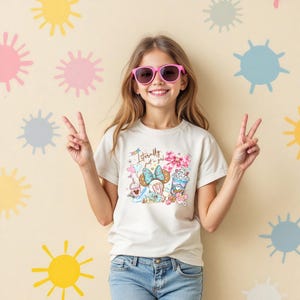 May include: A young person wearing pink sunglasses and a cream-colored t-shirt with the text "Literally just a girl." The shirt features a colorful graphic of a bow, desserts, and other cute illustrations. The background is a light beige with colorful shapes.